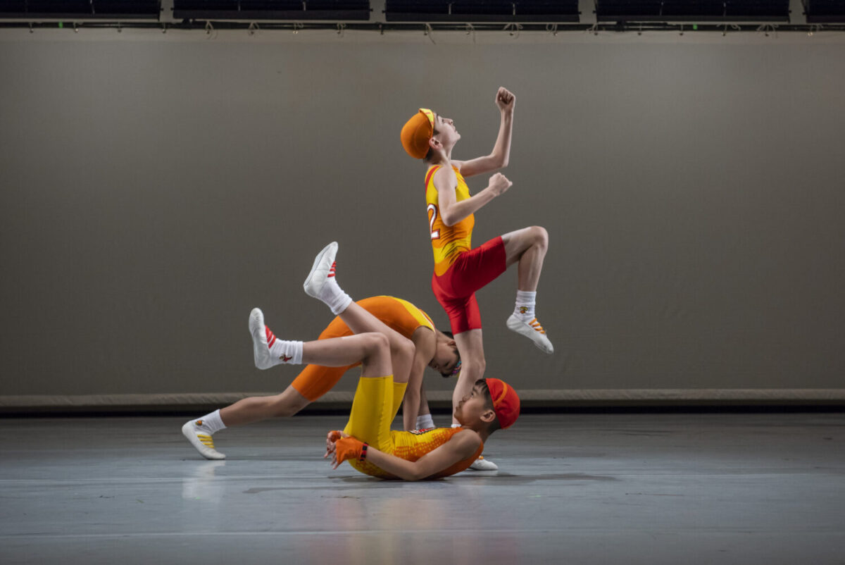 Three performers dressed in colorful athletic outfits are engaged in a dynamic, athletic dance routine. Two are low to the ground, while the third is wearing a cap and is posed as if mid-run, creating a visually striking tableau against a plain backdrop.